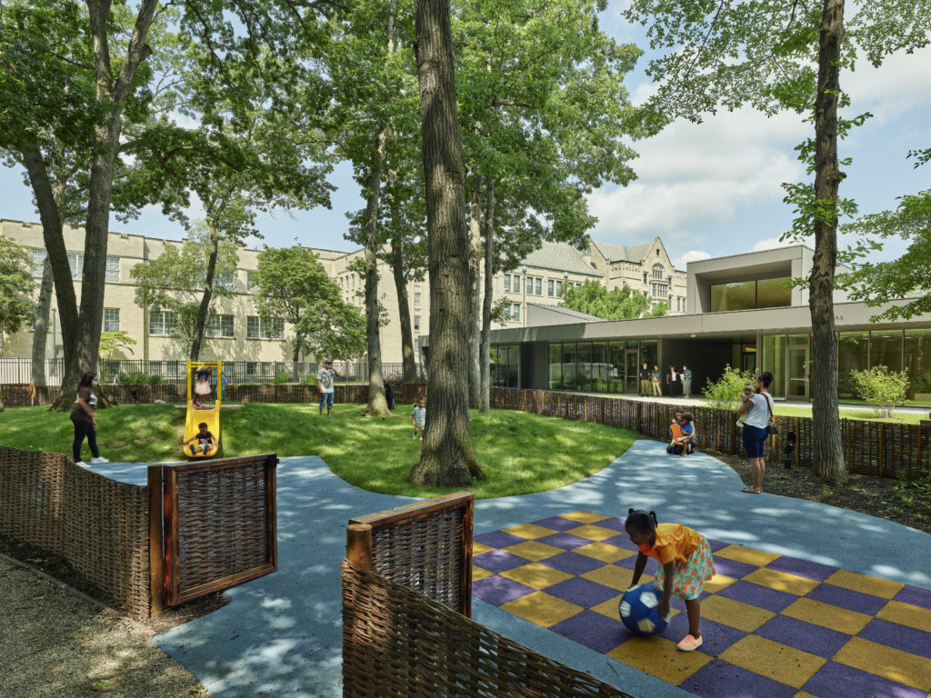 A playground with trees, a grassy area, blue pathways, and a checkered play area. A modern building is visible in the background.