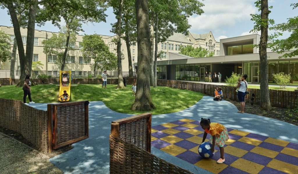 A playground with trees, a grassy area, blue pathways, and a checkered play area. A modern building is visible in the background.