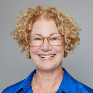 A white woman with short blonde curly hair and glasses is facing the camera and smiling. She is wearing a blue shirt. The background is gray.