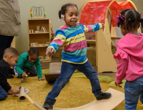 Children playing at an ECE center in Detroit. One child never the center is balancing, with arms outstretched, on a board curved like the foot of a rocking horse.