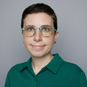 A white person with short brown hair, glasses, earrings, and a green top is facing the camera and smiling. The background is gray. - Alithea Tashey