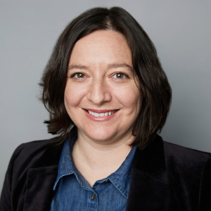 A white woman with short brown hair is facing the camera and smiling. She is wearing a blue denim shirt and black blazer. The background is gray. - Jessica Nepomiachi