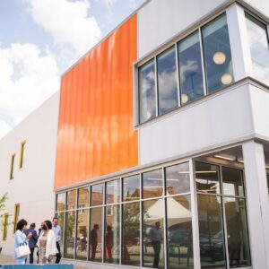 Exterior of a two-story, orange and white building at a city block corner, with tall windows, people chatting together out front, and someone entering the front door