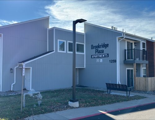 Apartment building with gray wood siding and red brick façade, featuring small balconies and an irregular roofline.