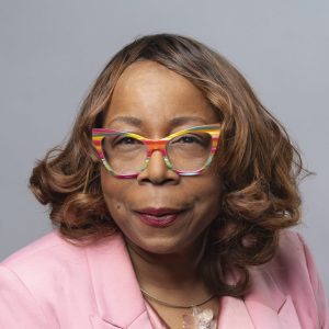 An African American woman with short brown hair and multicolored glasses is facing the camera and smiling. She is wearing a pink blazer. The background is gray. - Celina Byrd