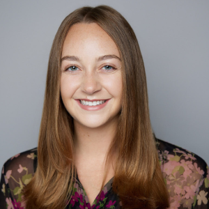 A white woman with straight long blonde hair is facing the camera and smiling. She is wearing a black and pink floral printed shirt. The background is gray. - Lily Yalowitz