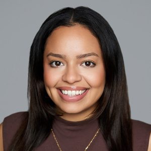 A Latina woman with straight black hair is facing the camera and smiling. She is wearing a brown top and a gold necklace. The background is gray. - Nathalie Gonzalez
