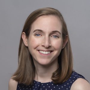 A white woman with straight brown hair is facing the camera and smiling. She is wearing a blue and white polka dotted top. The background is gray. - Kate Humphrey