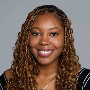 A Black woman with brown curly hair is facing the camera and smiling. She is wearing a black and white striped top. The background is gray. - A’Rya Pratt