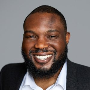 A Black man with short black hair and a black beard is facing the camera and smiling. He is wearing a white collared shirt and a black suit jacket. The background is gray. - Ola Odukoya