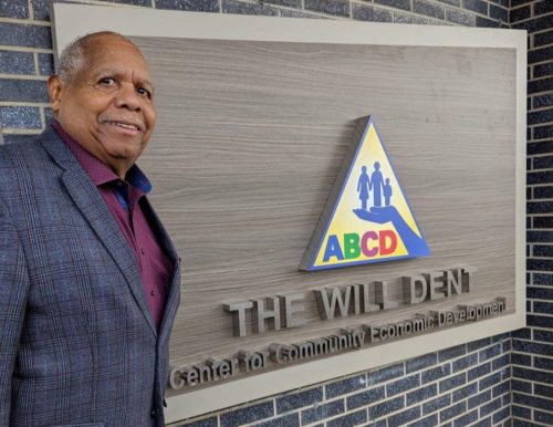 A person standing next to a wall sign attached to a building made of dark bricks. The person is wearing a checked jacket over a pink shirt. The sign is rectangular, featuring a triangular logo with stylized figures and colorful letters "ABCD" within it. Below the logo, there is text in large, raised letters that reads, "The Will Dent Center for Community Economic Development."