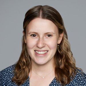 A white woman with shoulder length blonde hair is facing the camera and smiling. She is wearing a blue shirt with white polka dots. The background is gray. - Andie Linker
