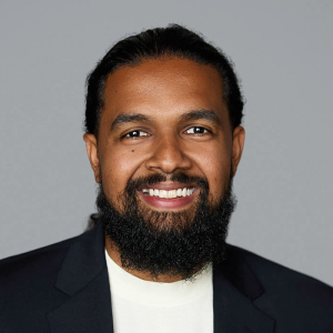 A Black man with short black hair and a black beard is facing the camera and smiling. He is wearing a white shirt and a black suit jacket. The background is gray. - Clay Kamaleson