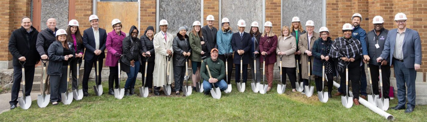 A group poses with shovels at a groundbreaking ceremony in front of a brick building with boarded windows. Many of the people in the group wear hard hats.
