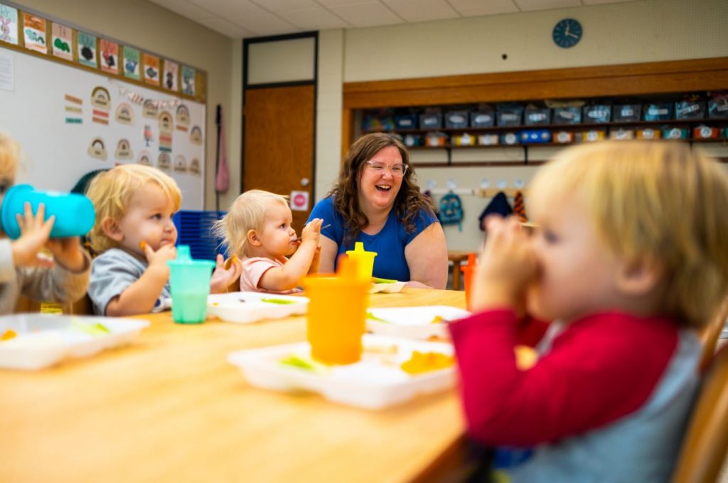 A smiling woman and several toddlers seated indoors at a table in a classroom engaged in learning activities, surrounded by educational materials.