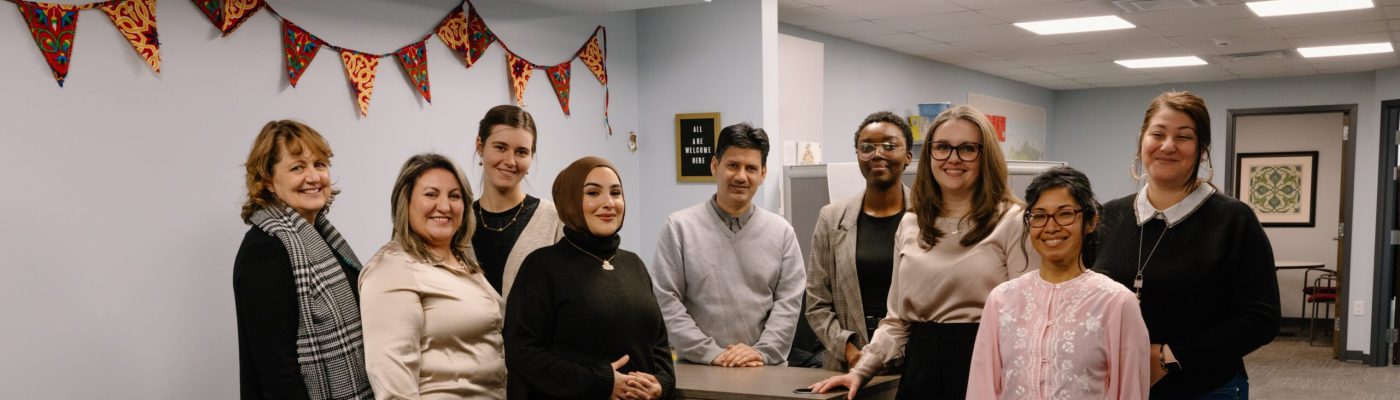 A group of people standing close together indoors, with a festive banner visible in the background. They are positioned around a counter in an office setting.