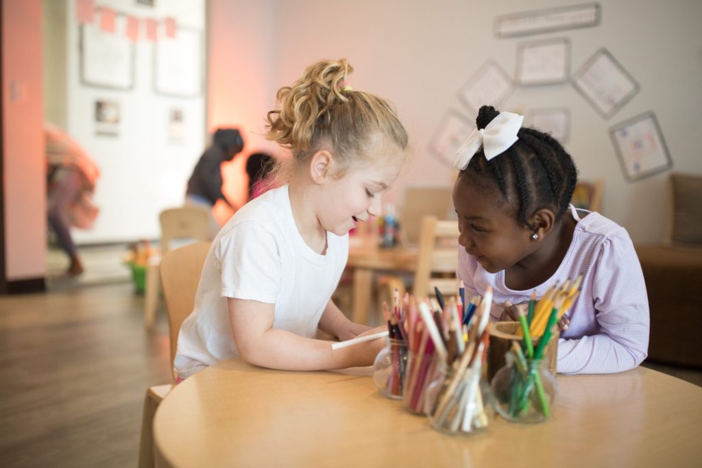 A girl sitting at a table, looking intently at her hands while another girl observes her. The setting is indoors with furniture visible and a wall in the background.