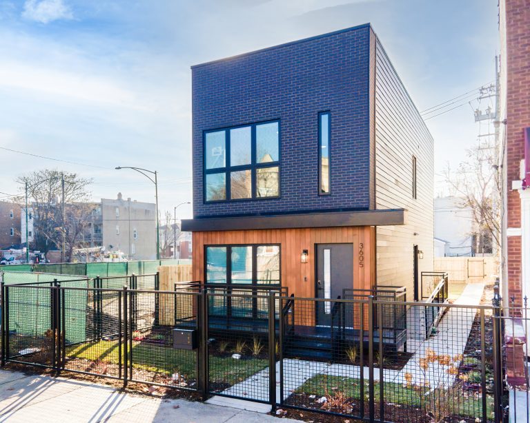 A modern two-story house with dark brick and wood paneling, large windows, and a black metal fence.