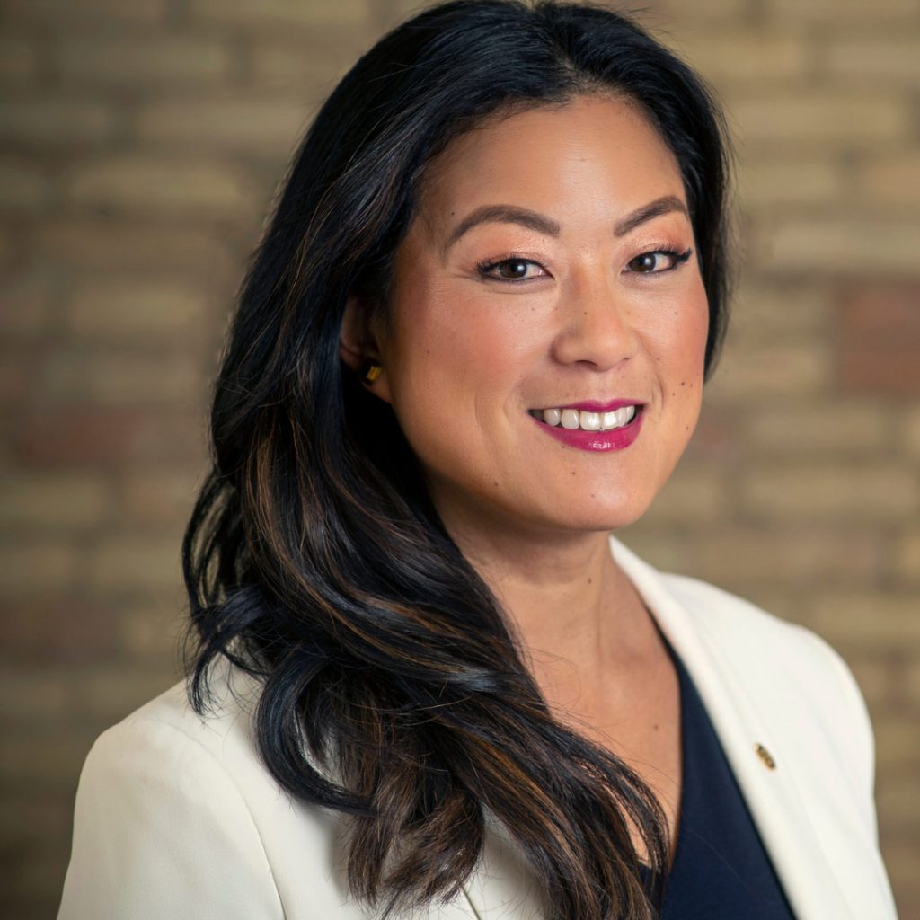 A woman with layered hair smiles at the camera. She is photographed in a headshot style against a brick wall background.