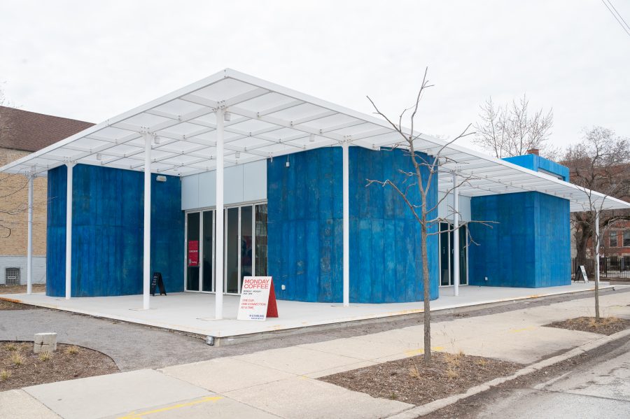 Modern building with bright blue walls and a white grid roof. A sidewalk leads to the entrance. A sign reads "Monday Coffee." Sparse trees in front.