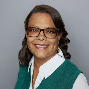A smiling woman wearing a white shirt with a green sweater in front of a grey backdrop. She is positioned in a headshot portrait style.