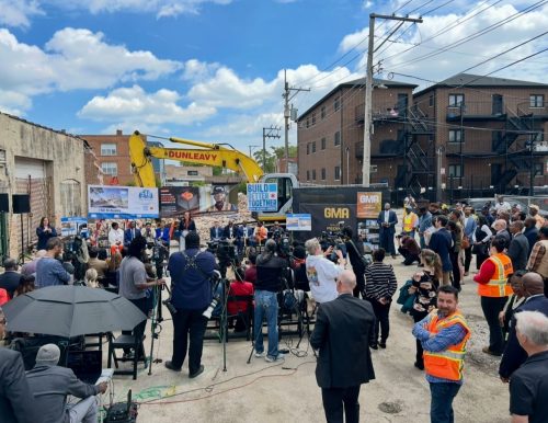 A large crowd gathers at an outdoor groundbreaking ceremony with speakers, cameras, and a yellow excavator in the background.
