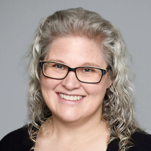 A white woman with curly silver hair is facing the camera and smiling. She is wearing glasses and a black top. The background is gray. - Michelle Cole
