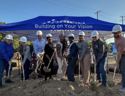 A group of people stand outside under a blue tent that says "Building on Your Vision." They are holding shovels standing in a pile of dirt.