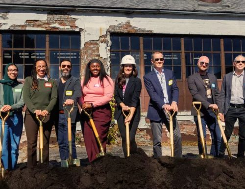 A group of ten people at a groundbreaking ceremony, each holding a golden shovel in front of an old building.