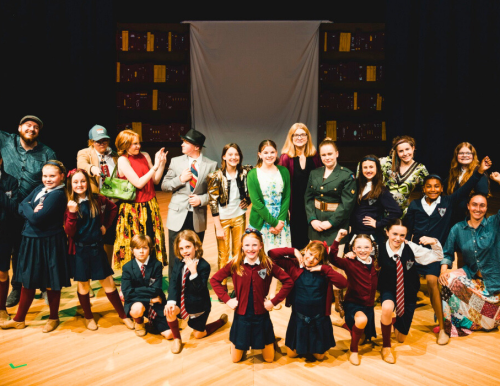 A group of children and adults on a stage, dressed in school uniforms and various costumes, with a theater backdrop.