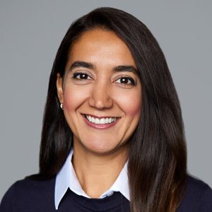 A woman with straight brown hair is facing the camera and smiling. She is wearing a white collared shirt under a navy sweater. The background is gray. - Golnaz Farzad