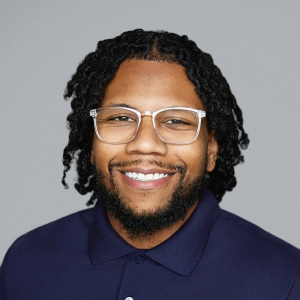A Black man with short black hair and a black beard is facing the camera and smiling. He is wearing glasses and a navy blue shirt. The background is gray. - Korrane Bateman