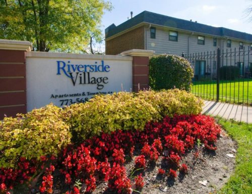 Entrance sign for Riverside Village apartments, with shrubs and a residential building in the background.