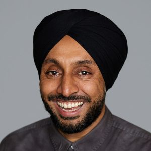 A man with short black facial hair is facing the camera and smiling. He is wearing a black turban and a dark gray top. The background is gray. - Sukh Kalsi