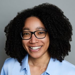An African American woman with curly brown hair and glasses is facing the camera and smiling. She is wearing a blue collared shirt. The background is gray. - Ashlyn Lee