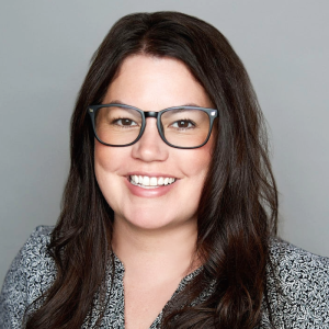 A white woman with straight brown hair is facing the camera and smiling. She is wearing glasses and a black and white top. The background is gray. - Aubrey Stevens
