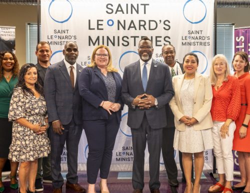 A diverse group of thirteen people posing in front of a "Saint Leonard's Ministries" banner.