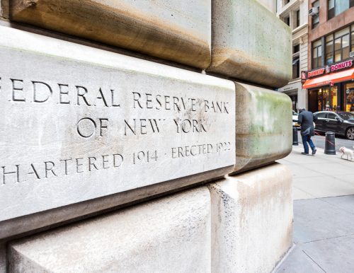Corner of a stone building engraved with text that says "Federal Reserve Bank of New York" beside a busy urban street with pedestrians and a Dunkin' Donuts storefront.