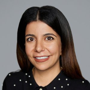 A white woman with straight brown hair is facing the camera and smiling. She is wearing a black top with white polka dots. The background is gray. - Hala Rousan