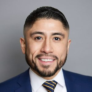 A Latino man with short brown hair and short brown facial hair is facing the camera and smiling. He is wearing a white shirt, striped tie, and blue suit jacket. The background is gray. - Jessi Ventura