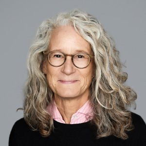 A white woman with curly silver hair is facing the camera and smiling. She is wearing glasses, a pink collared shirt, and a black sweater. The background is gray. - Lynn Carpenter