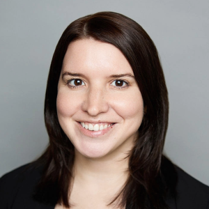 A white woman with straight brown hair is facing the camera and smiling. She is wearing a black blazer. The background is gray. - Megan Adamczewski