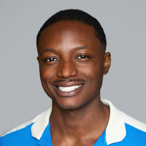 A Black man with short black hair is facing the camera and smiling. He is wearing a blue and white shirt. The background is gray. - DaMarcus Lacy