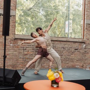 Two people wearing beige outfits dance on a stage in front of a brick wall with a big square window