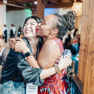 A woman embraces another woman and kisses her cheek, both smiling, in a room full of people