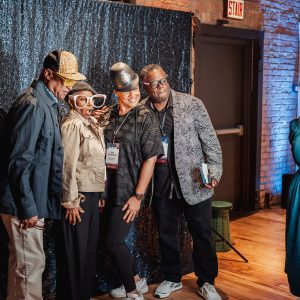 Four people in silly hats stand in front of a sequin background while getting their picture taken by a photo booth