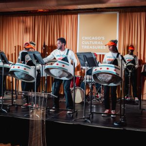 A group of young people perform on steel drums on a stage in front of a yellow curtain and a square sign that reads 