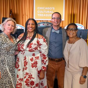 Four people stand smiling in front of a stage and a yellow curtain and a square sign that reads 