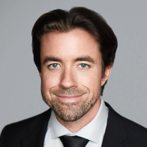 A white man with wavy brown hair and short brown facial hair is facing the camera and smiling. He is wearing a white collared shirt, black tie, and black suit jacket. The background is gray. - Lucas Hopkins
