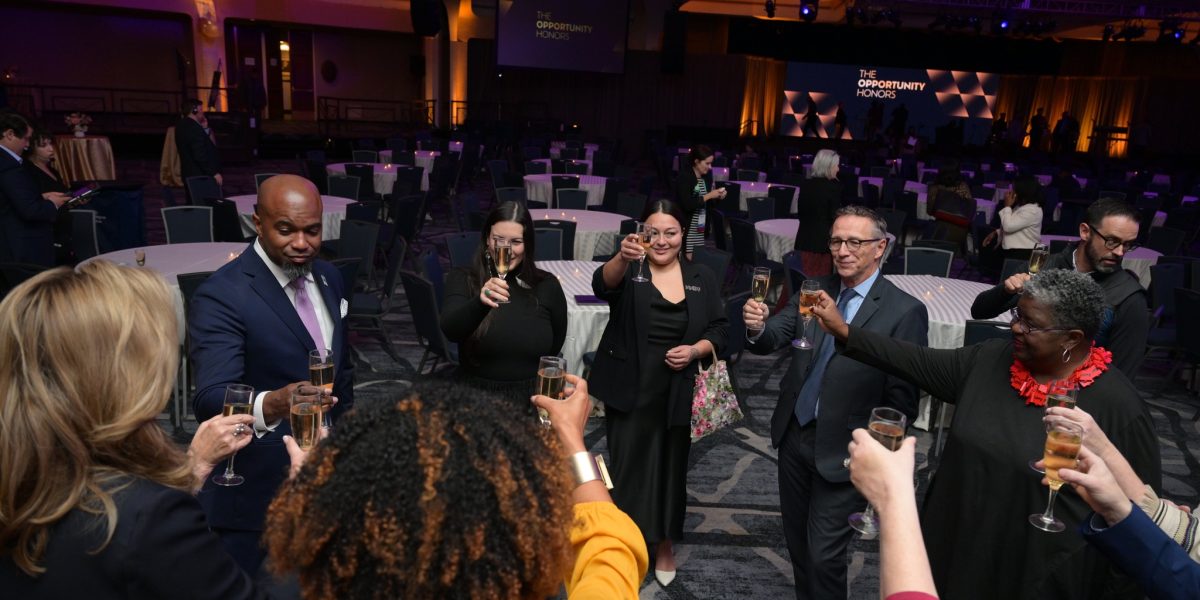 The image shows a group of people standing in a circle, raising glasses in a toast. They are in a large, dimly lit ballroom with round tables and chairs arranged throughout the room. The lighting is a mix of warm and purple hues, highlighting the ceiling's arches and creating a festive atmosphere. In the background, there is a stage with a large screen displaying the text 